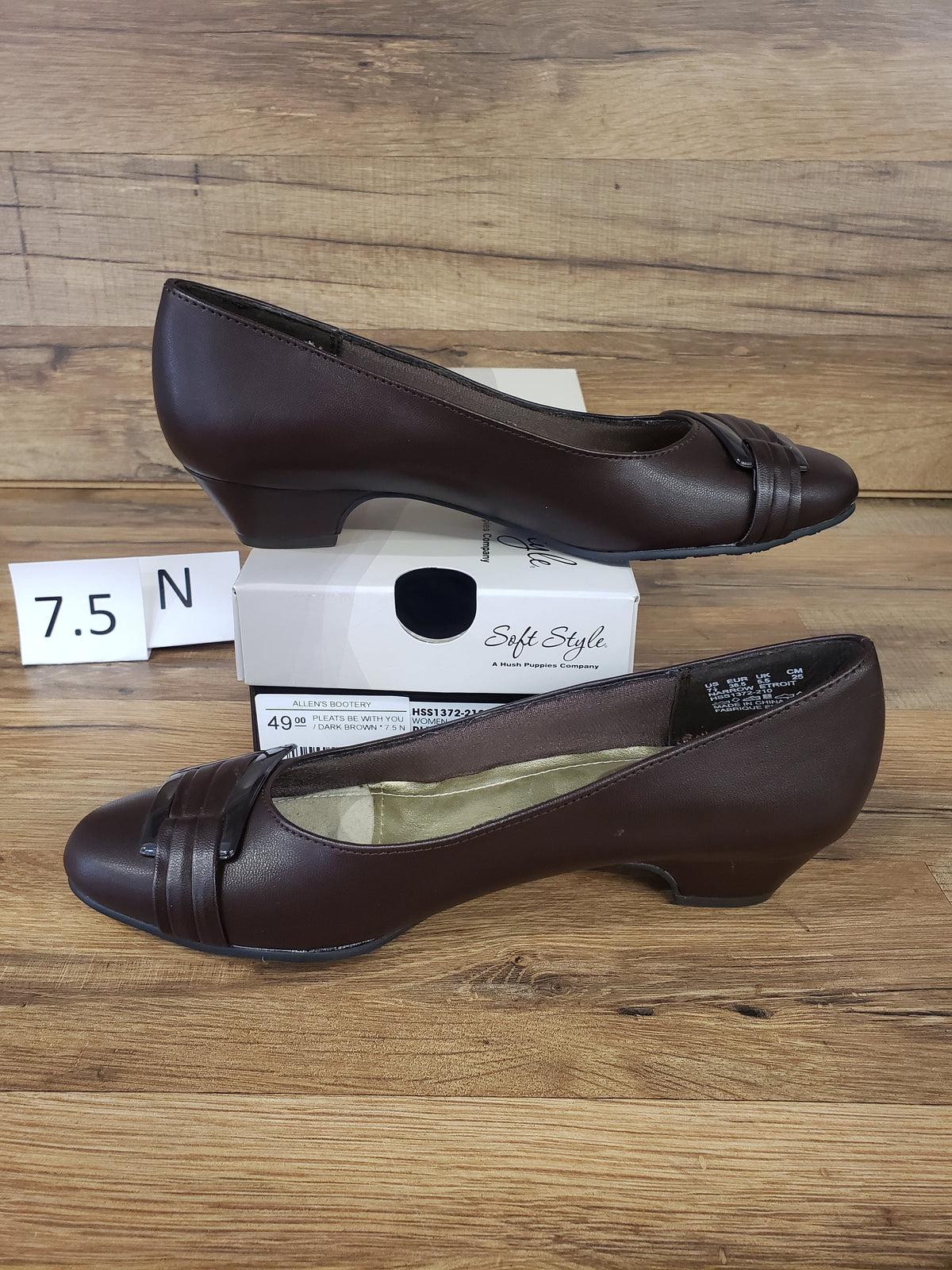 Allen's Bootery dark brown closed-toe pumps with pleated toe detail, buckle accent, and low heel on a wooden surface.