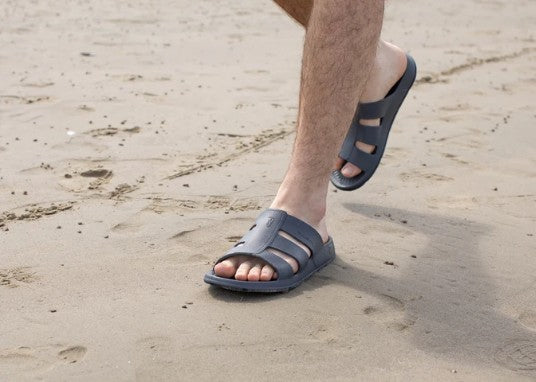 Allen's Bootery black Stanley Slide sandals worn by a man walking on a sandy beach for comfort and water activities.