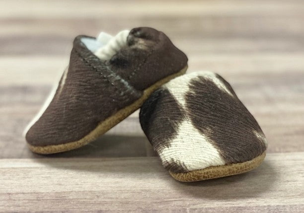 Allen's Bootery soft sole infant moccasins featuring a brown and white cow print pattern on a wooden surface.