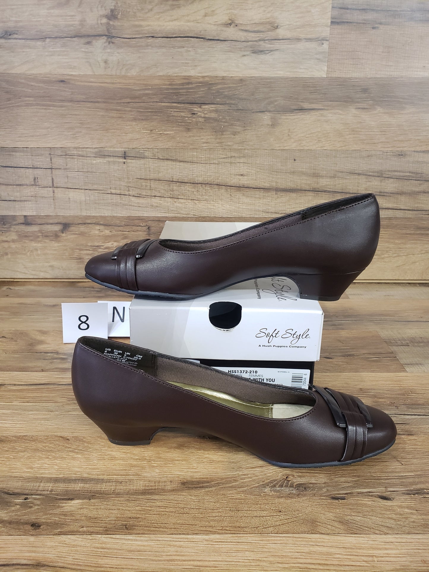 Allen's Bootery dark brown closed-toe pumps with pleated buckle detail, rounded toe, and low heel on a wooden surface.