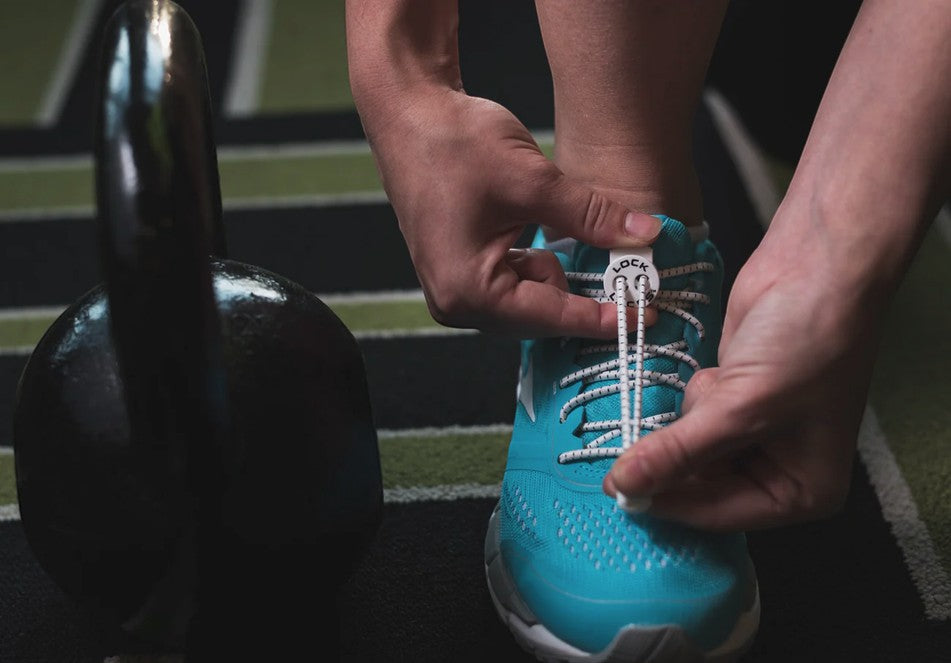 Allen's Bootery white LOCK LACES no-tie shoelaces being adjusted on a bright blue running shoe near a kettlebell