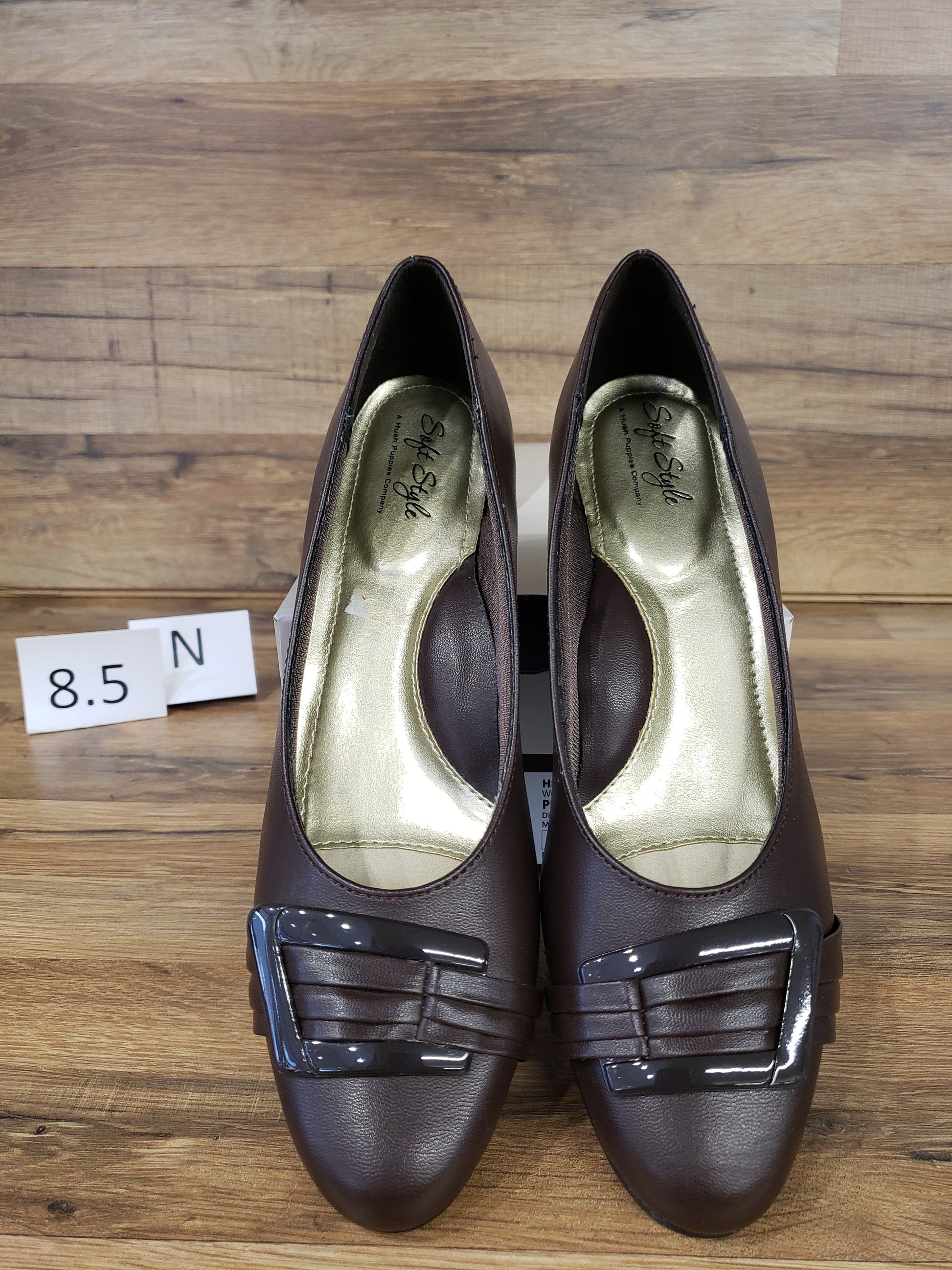 Allen's Bootery dark brown closed-toe pumps with a decorative buckle and pleat detail on a wooden background.