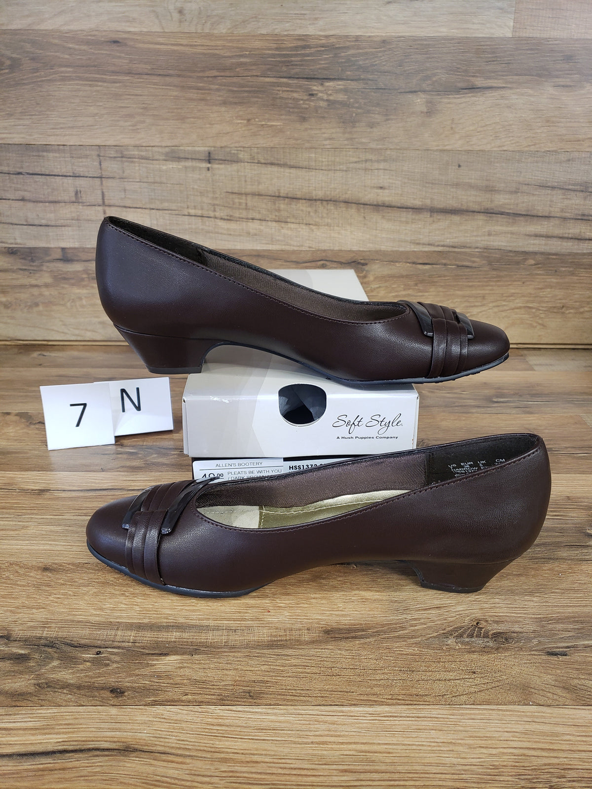 Allen's Bootery dark brown closed-toe pumps with pleated buckle detail, rounded toe, and low heel on a wooden surface.