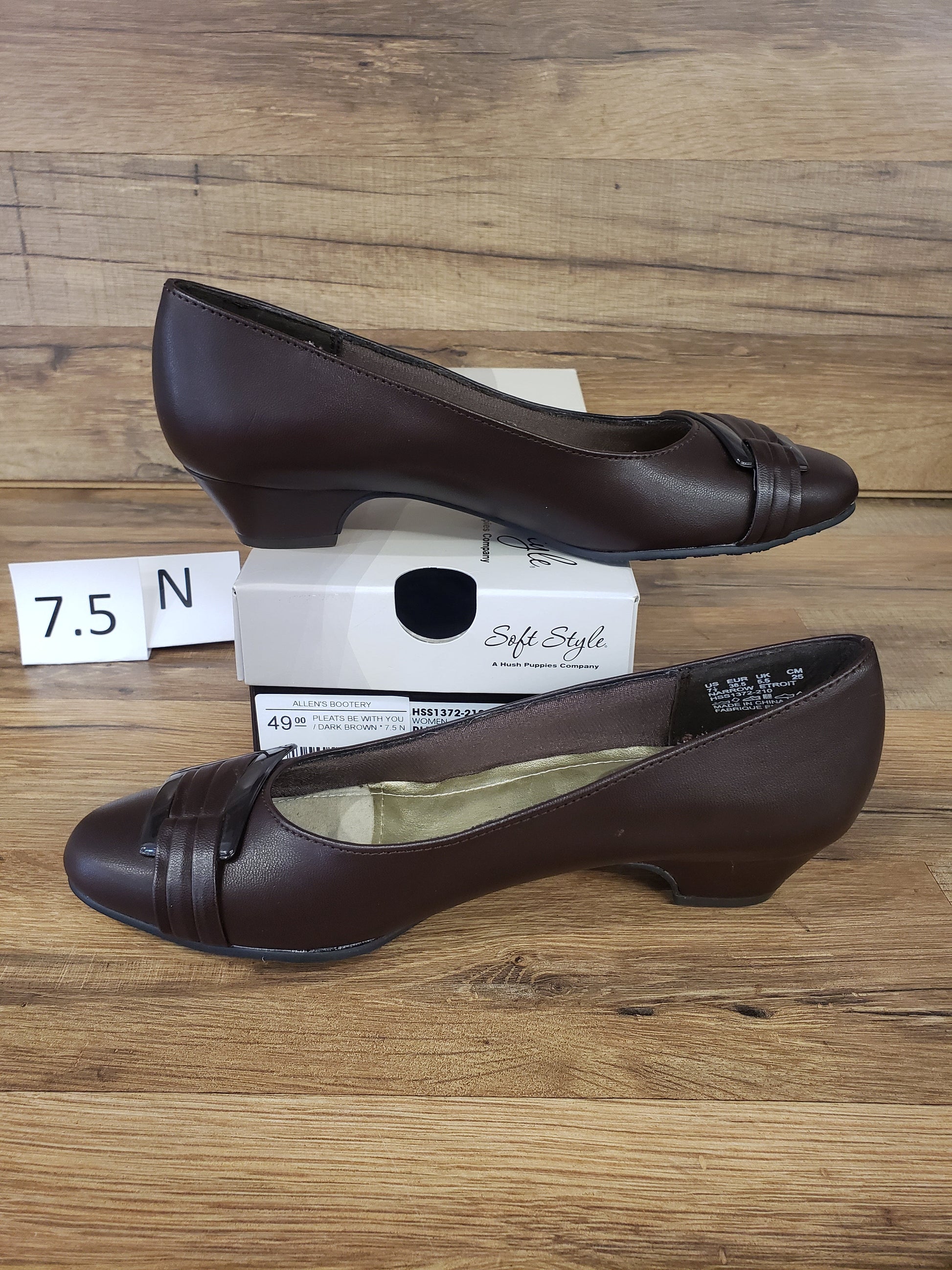 Allen's Bootery dark brown closed-toe pumps with pleated toe detail, buckle accent, and low heel on a wooden surface.