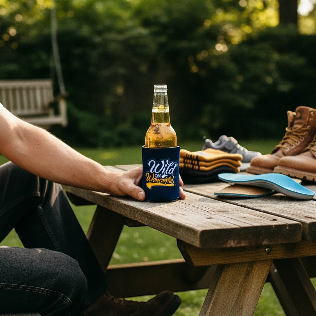 Allen's Bootery navy blue Wild & Wonderful patch can cooler holding a glass beer bottle on an outdoor wooden table.