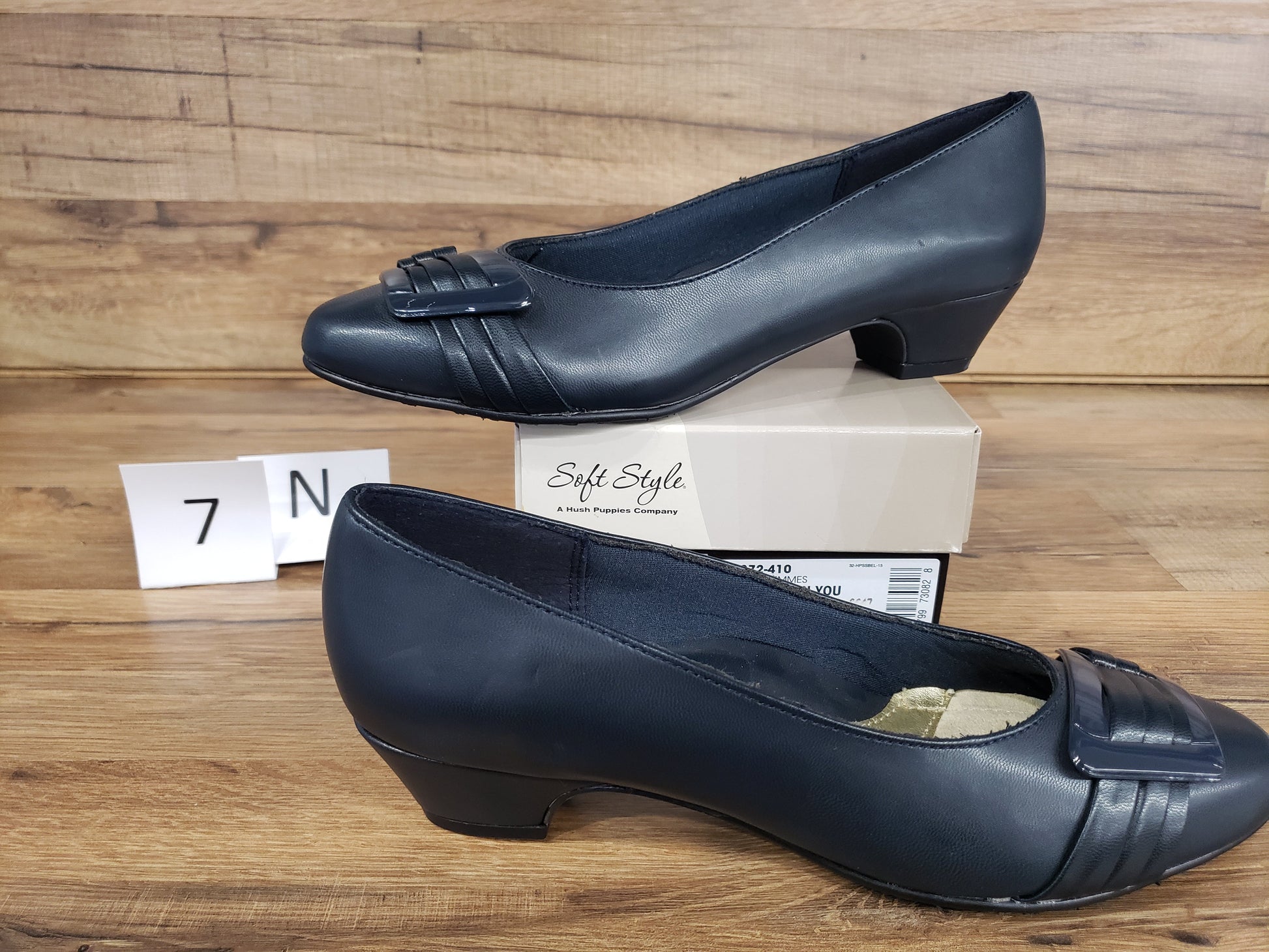 Allen's Bootery navy blue closed-toe pumps with pleated toe detail, buckle ornament, and low heel on a wooden surface.