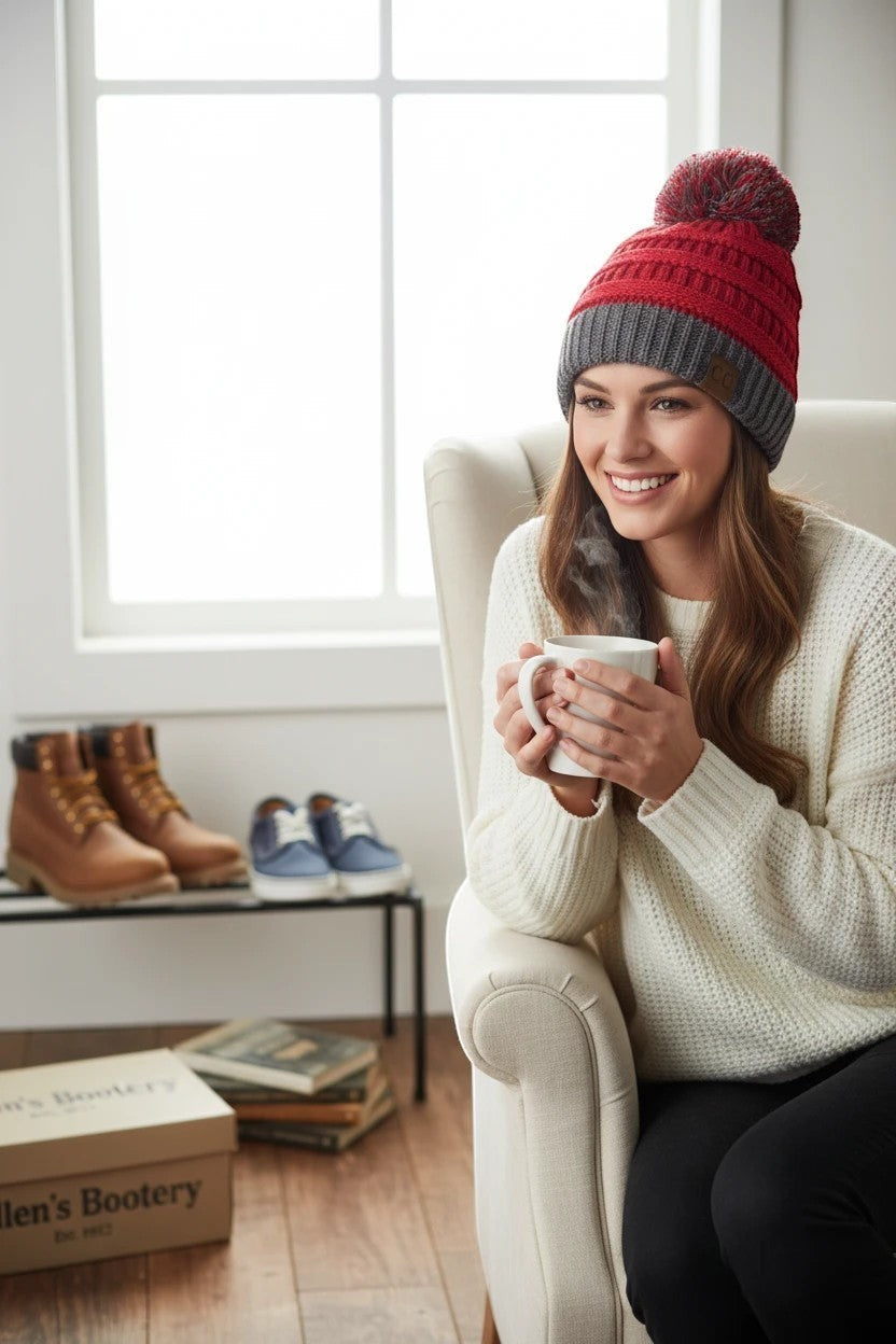 Woman wearing an Allen's Bootery red and charcoal grey cable-knit pom-pom beanie while holding a steaming mug.